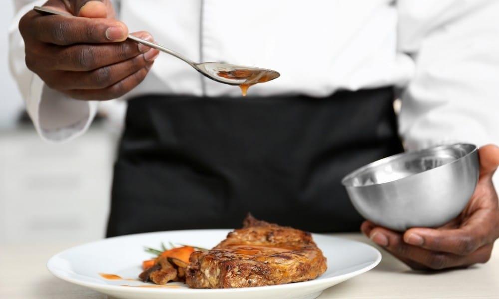 A chef is pouring a thick brown sauce from a silver ladle onto a steak on a white plate.