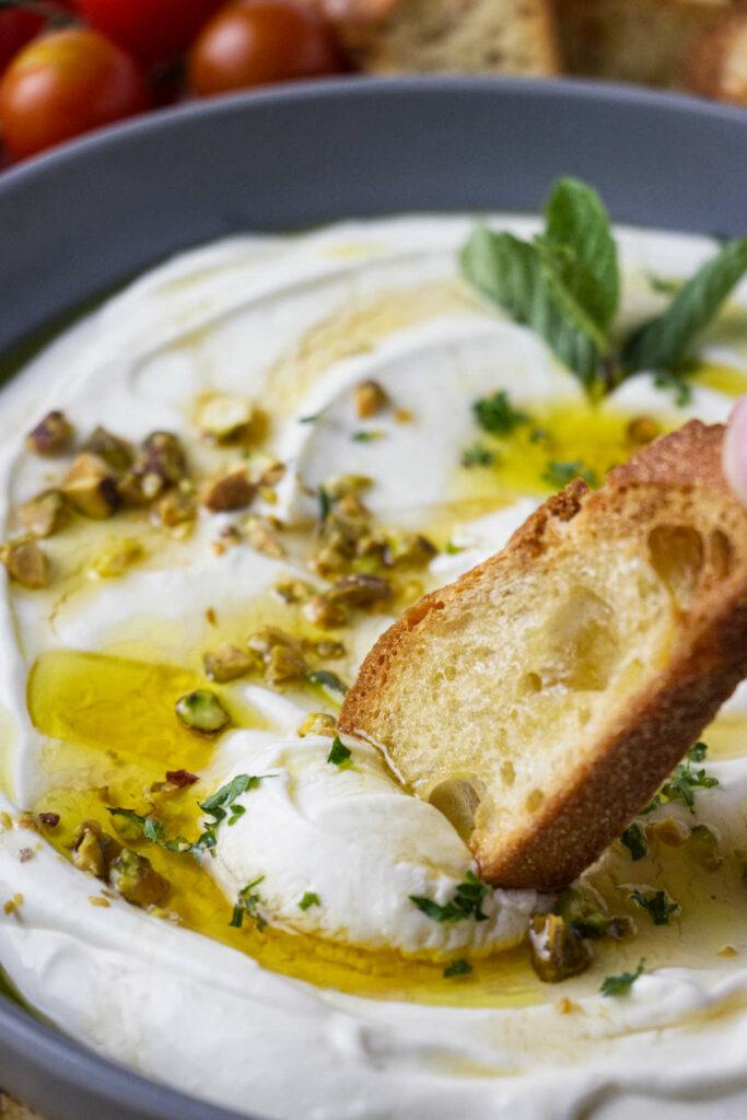 A piece of bread is dipped in a bowl of labneh, which is topped with olive oil, pistachios, and mint.