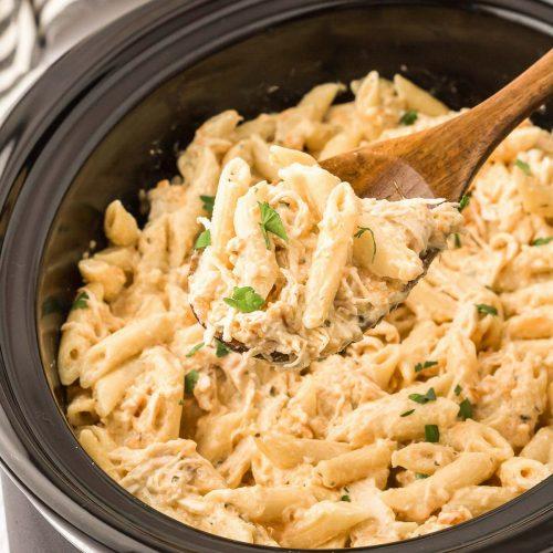 A wooden spoon is lifting creamy chicken pasta out of a slow cooker.